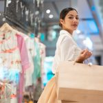 Young Asian woman holding shopping bag and looking at shop window while walking in shopping mall